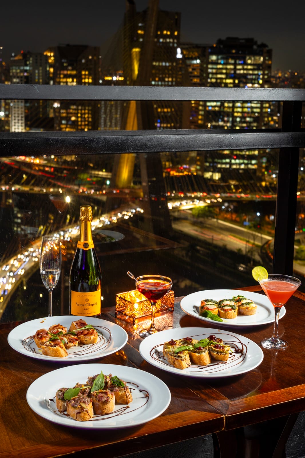 Bruschettas, espumante e coquetéis servidos na varanda com vista noturna para a ponte estaiada